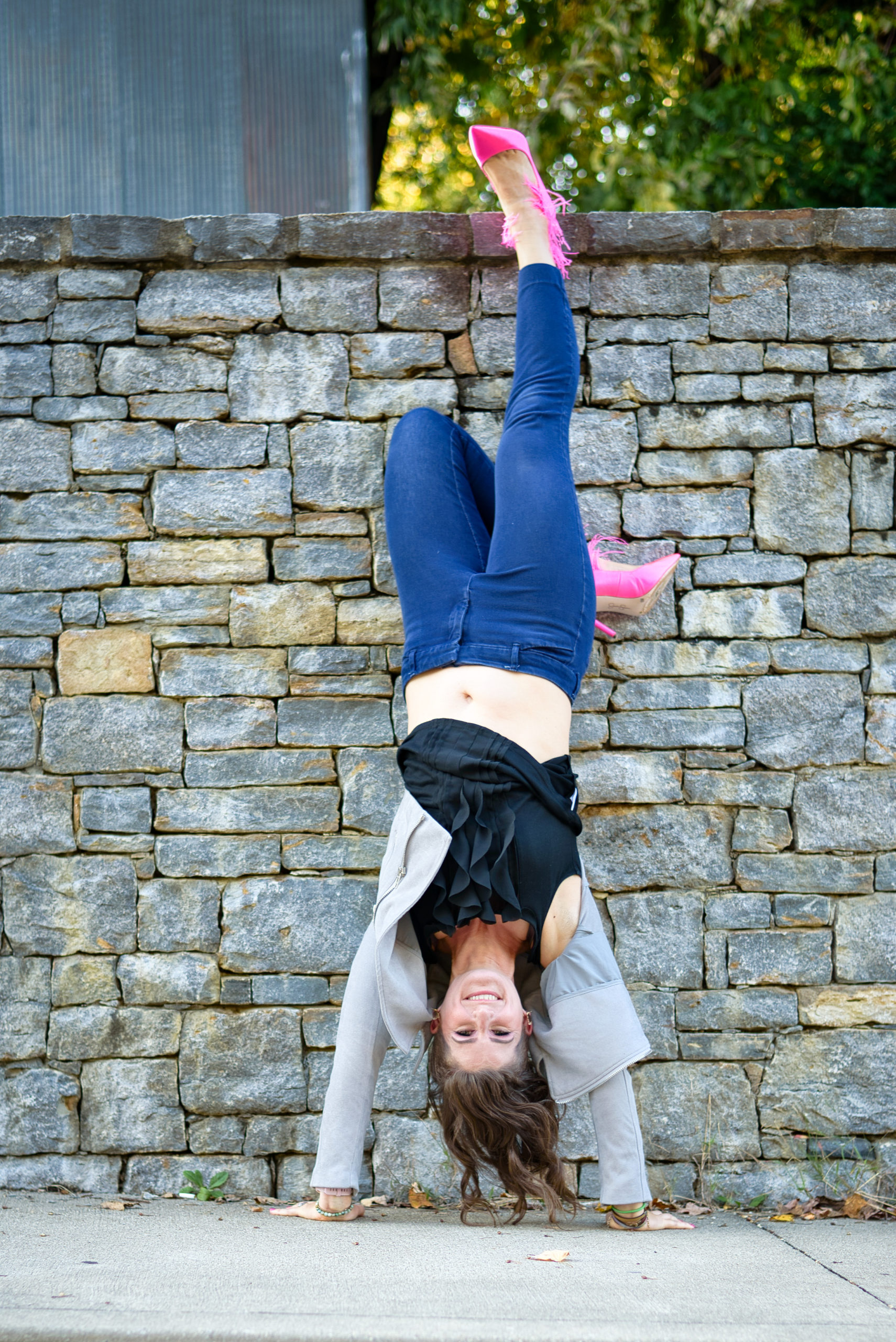 Dr. Andrea Moore does a hand stand in front of a stone wall while wearing hot pink high heels. She is promoting her Pain to Power Group for chronic pain.